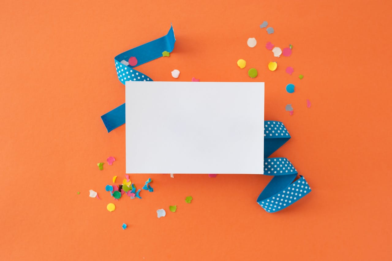 Flat lay of colorful confetti and blue ribbon with blank card on orange background. Perfect for celebrations.